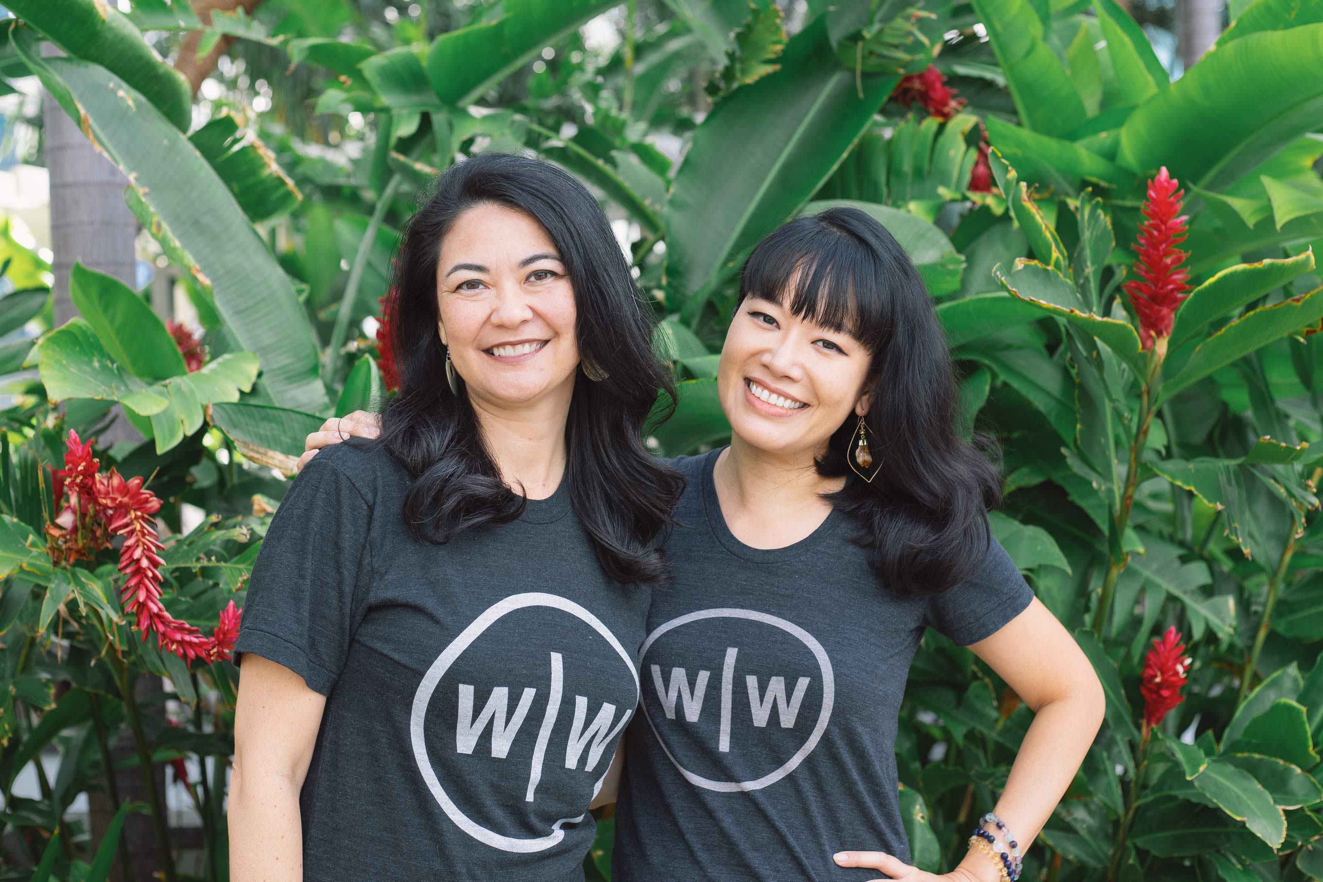 Malia Wisch and Jane Nguyen smiling with Wall-to-Wall logo shirts in a tropical, Hawaii setting